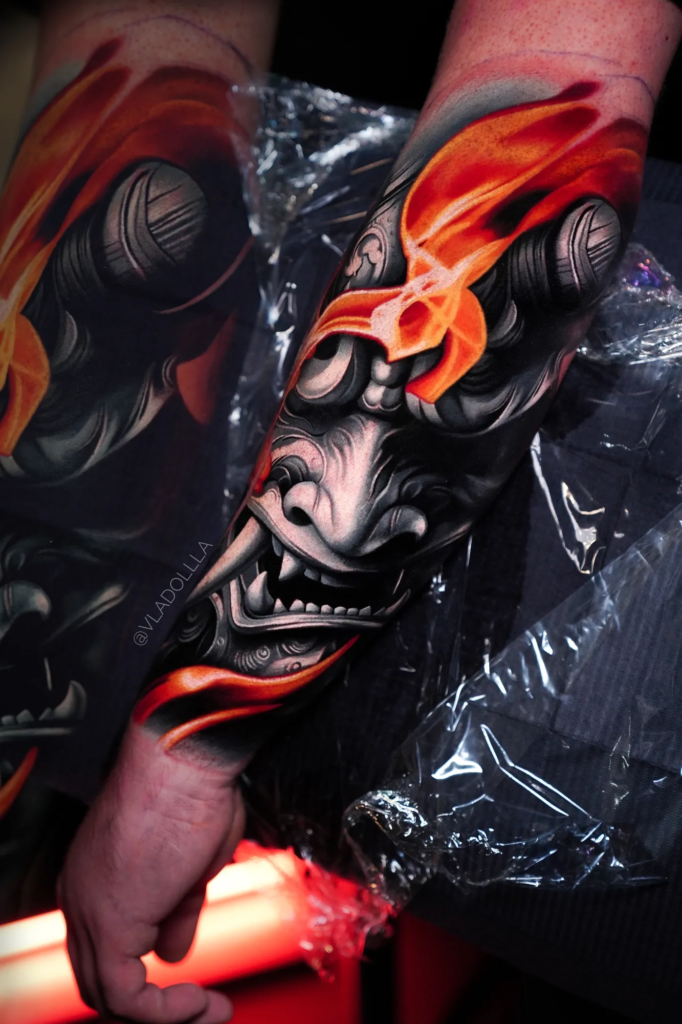 Forearm tattoo of a grey hannya mask with orange flames across its brow, fresh under plastic wrap.