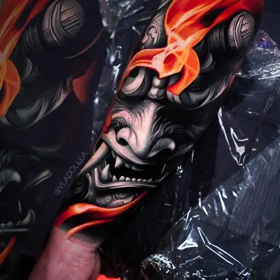 Forearm tattoo of a grey hannya mask with orange flames across its brow, fresh under plastic wrap.