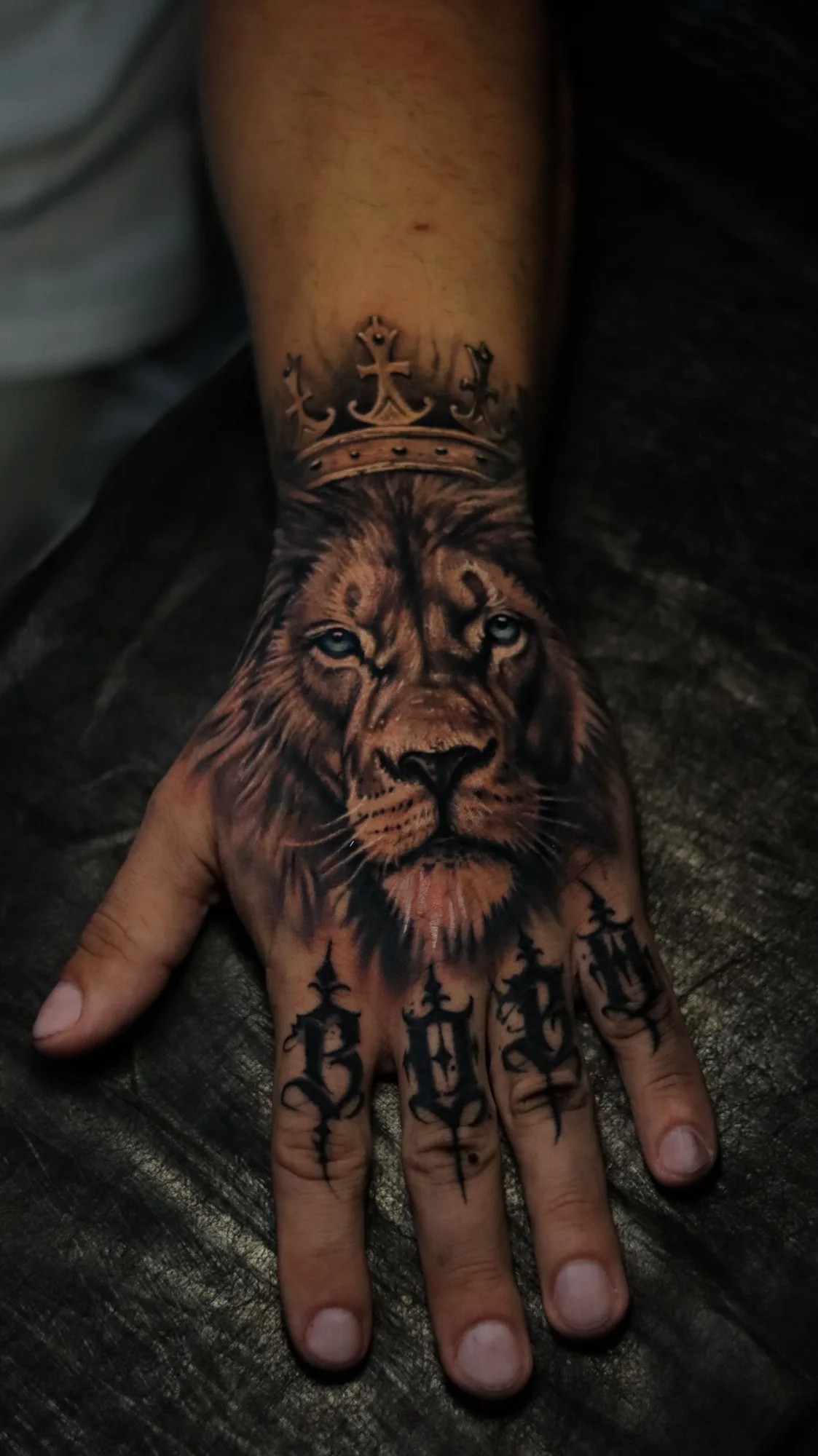 Black and grey realism tattoo on the back of the hand: a lion's face with pale blue eyes under a crown, with gothic lettering across the knuckles.