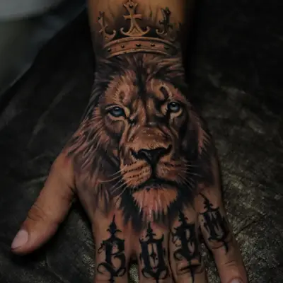Black and grey realism tattoo on the back of the hand: a lion's face with pale blue eyes under a crown, with gothic lettering across the knuckles.