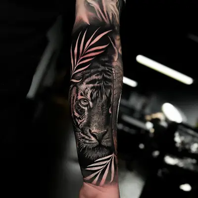 Black and grey realism tattoo on the inner forearm: a tiger's face framed by light palm fronds sweeping in from above and below.