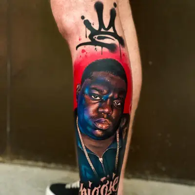 Color realism portrait tattoo on the calf: The Notorious B.I.G. in blues and purples against a red backdrop, with a dripping graffiti crown above and "Biggie" in script below.