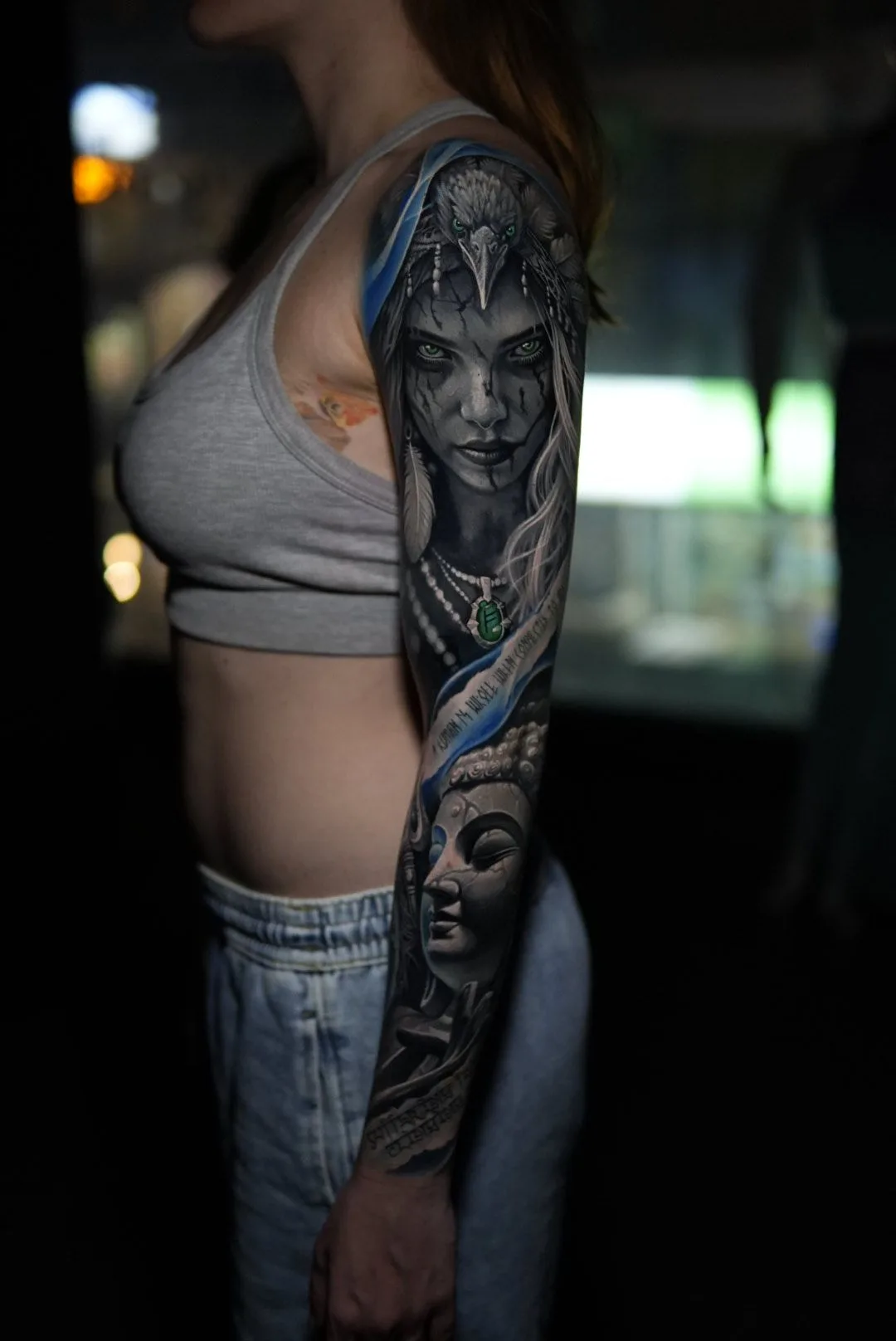 Black and grey full sleeve with electric blue accents: a woman with dark face paint wearing a bird headdress on the upper arm, and a carved stone face lower on the forearm.