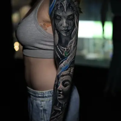 Black and grey full sleeve with electric blue accents: a woman with dark face paint wearing a bird headdress on the upper arm, and a carved stone face lower on the forearm.