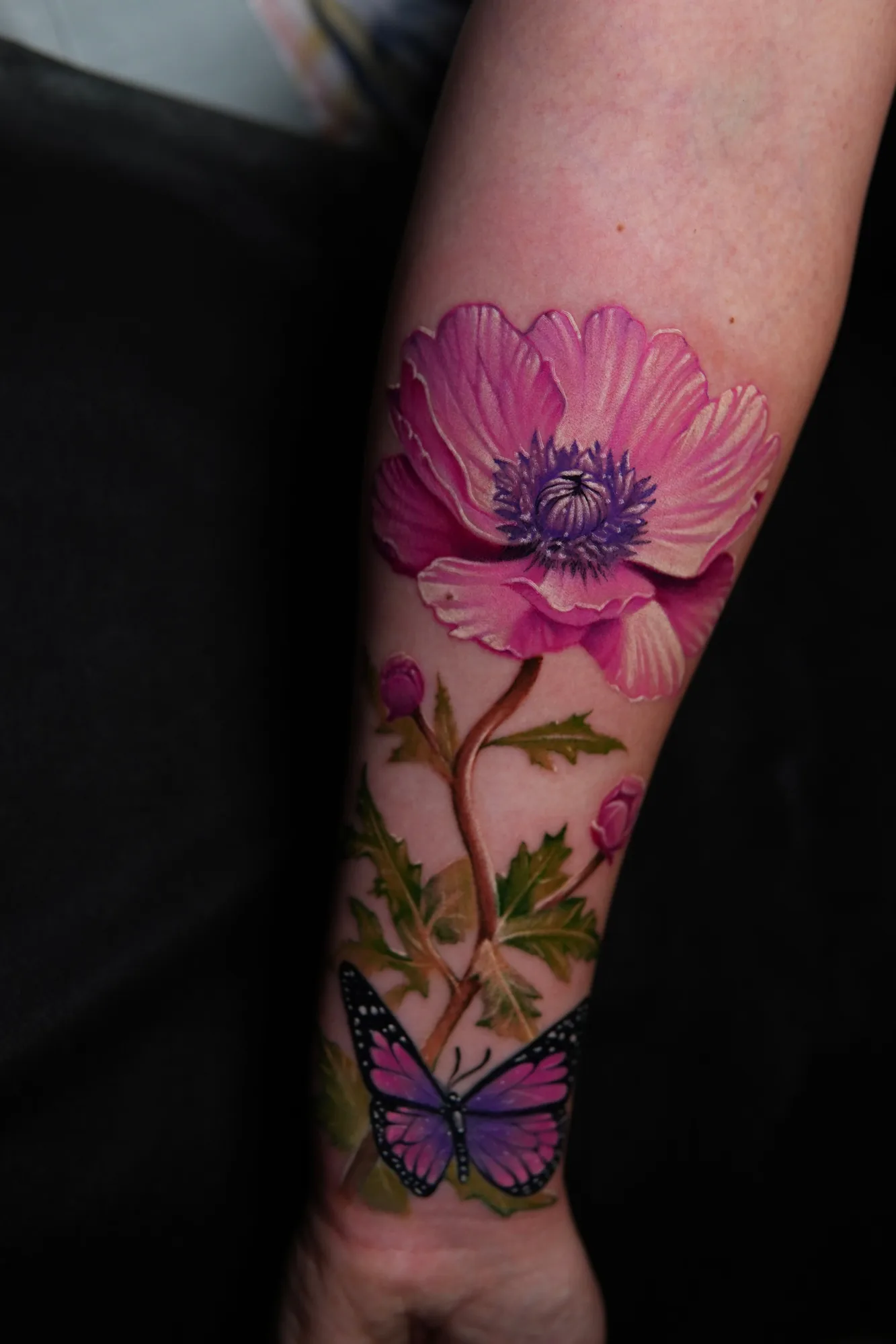 Color realism tattoo on the forearm: a large pink flower with a purple center, its stem and leaves running down toward the wrist to a small purple butterfly.