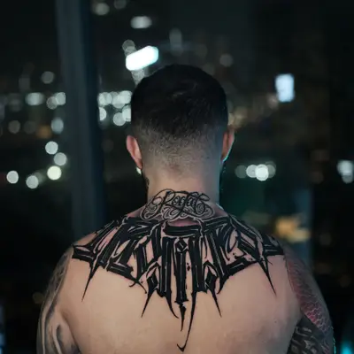 Black calligraphy tattoo across the upper back: a wide word in sharp, heavy letterforms with a smaller script above it at the base of the neck.
