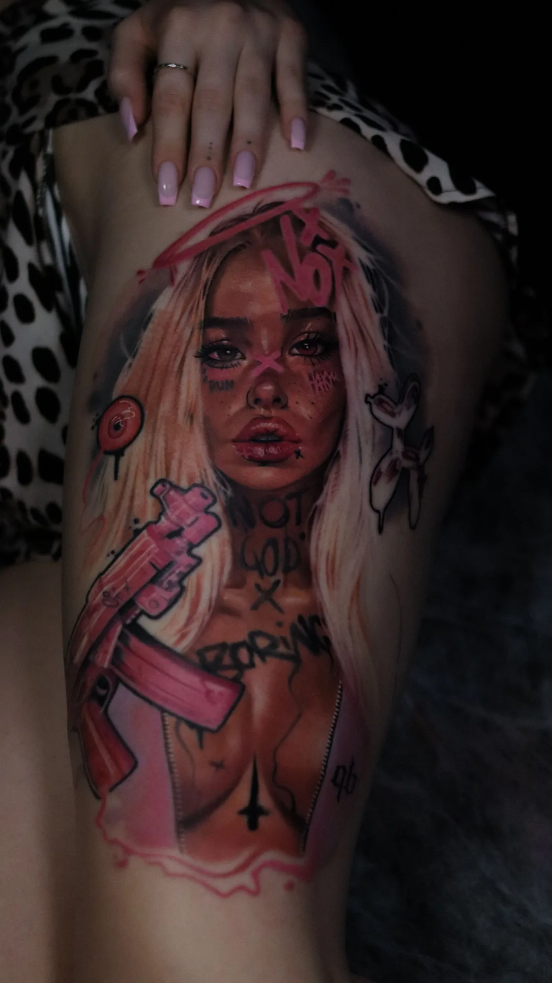 Color realism tattoo on the thigh: a blonde woman with a pink halo holding a pink rifle, with hot pink graffiti marks across her face and body.