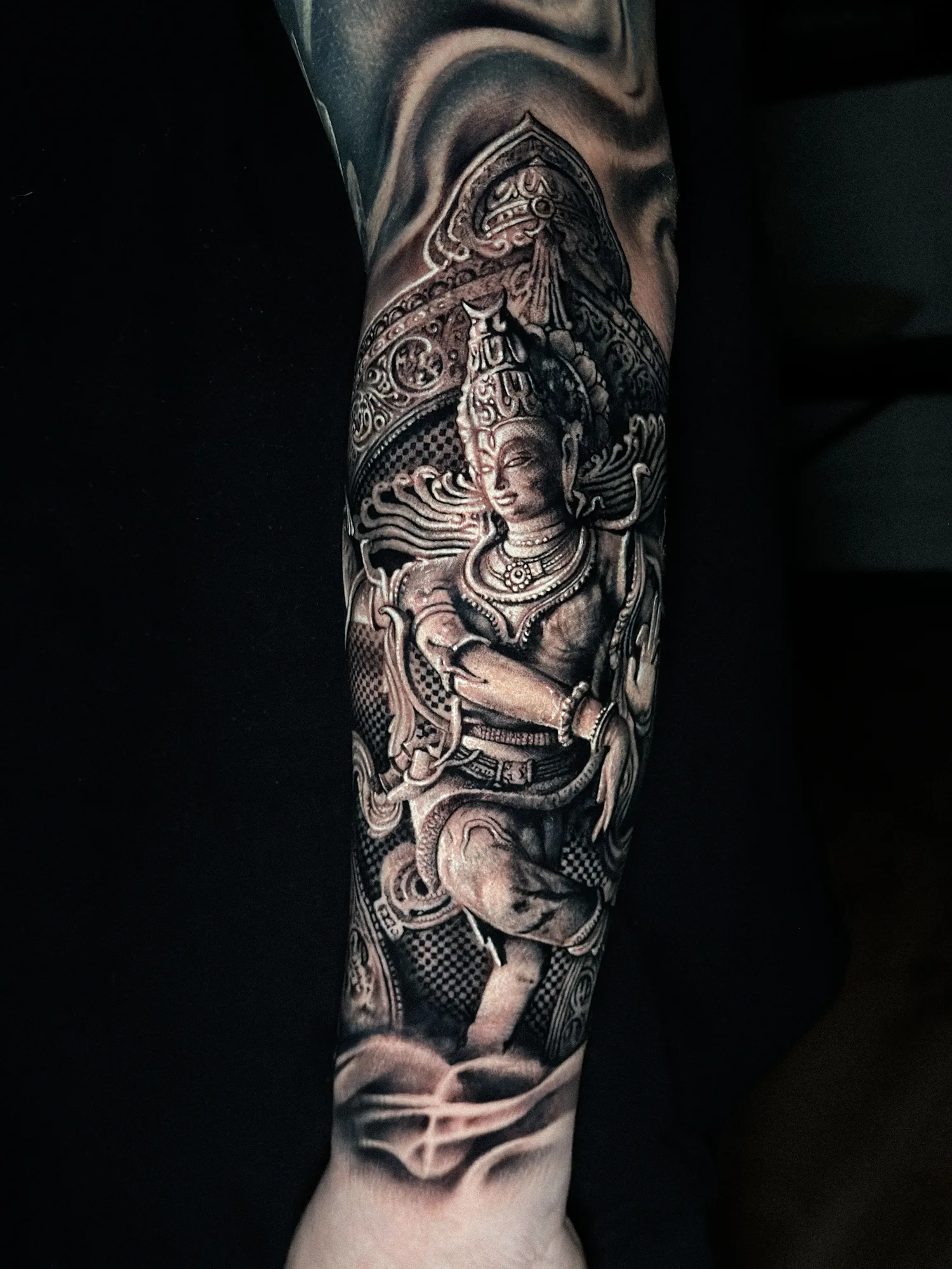 Black and grey realism tattoo on the forearm: a carved stone deity figure inside an ornate temple arch, fading into smoke at the top and bottom.