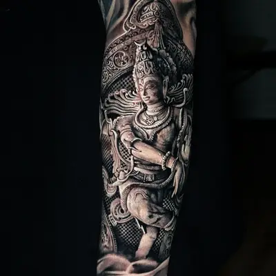 Black and grey realism tattoo on the forearm: a carved stone deity figure inside an ornate temple arch, fading into smoke at the top and bottom.