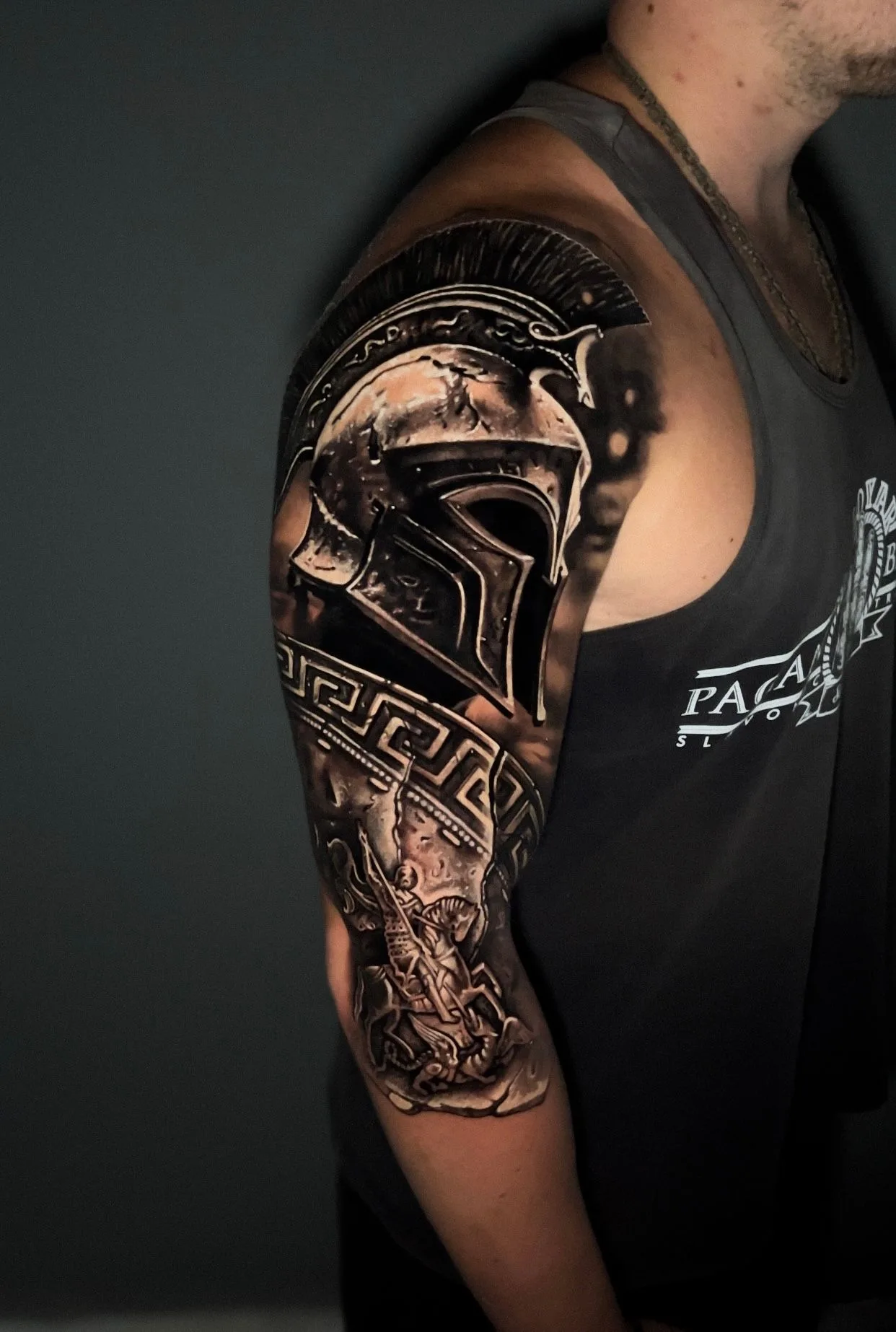 Black and grey realism half sleeve on the upper arm: a polished Spartan helmet on the shoulder above a Greek key band, with a warrior scene below.