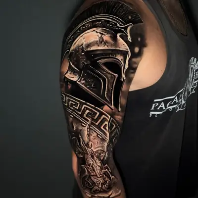 Black and grey realism half sleeve on the upper arm: a polished Spartan helmet on the shoulder above a Greek key band, with a warrior scene below.