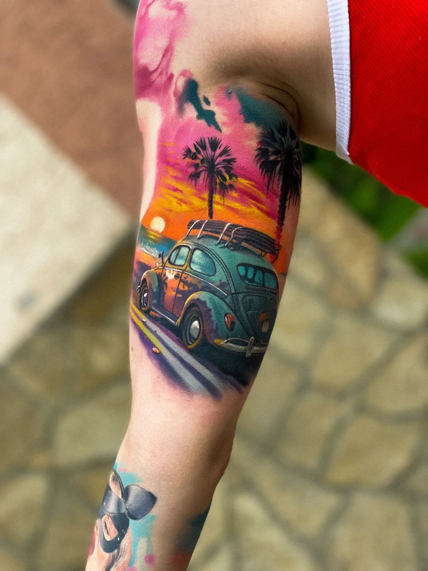 Color tattoo on the inner upper arm: a vintage Volkswagen Beetle with a surfboard on the roof driving past palm trees into a pink and orange sunset, with watercolor splashes at the edges.