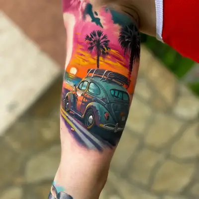 Color tattoo on the inner upper arm: a vintage Volkswagen Beetle with a surfboard on the roof driving past palm trees into a pink and orange sunset, with watercolor splashes at the edges.