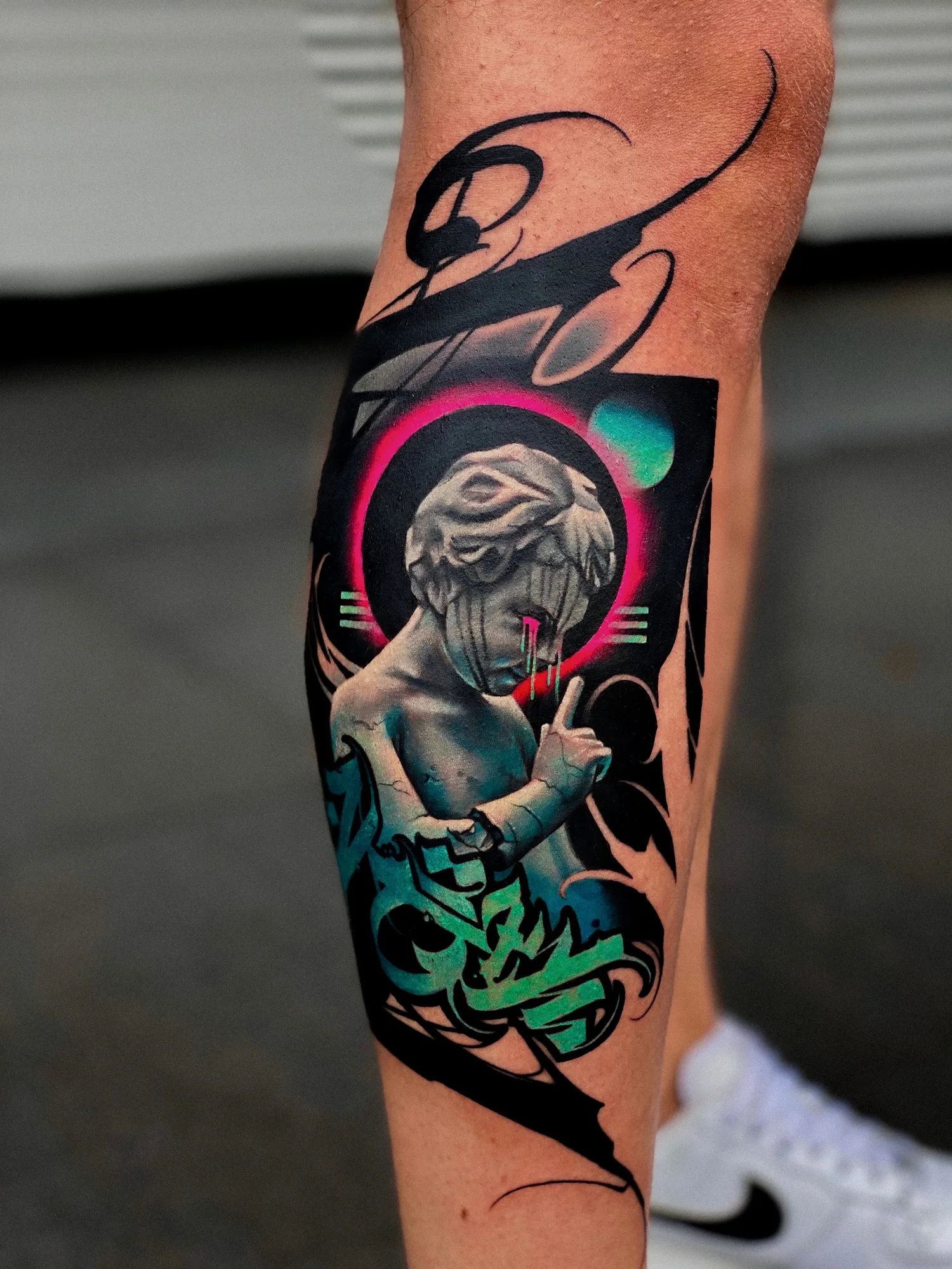 Tattoo on the calf: a grey marble cherub raising a finger to its lips inside a pink ring, with teal graffiti lettering below and sweeping black brushstroke calligraphy around it.
