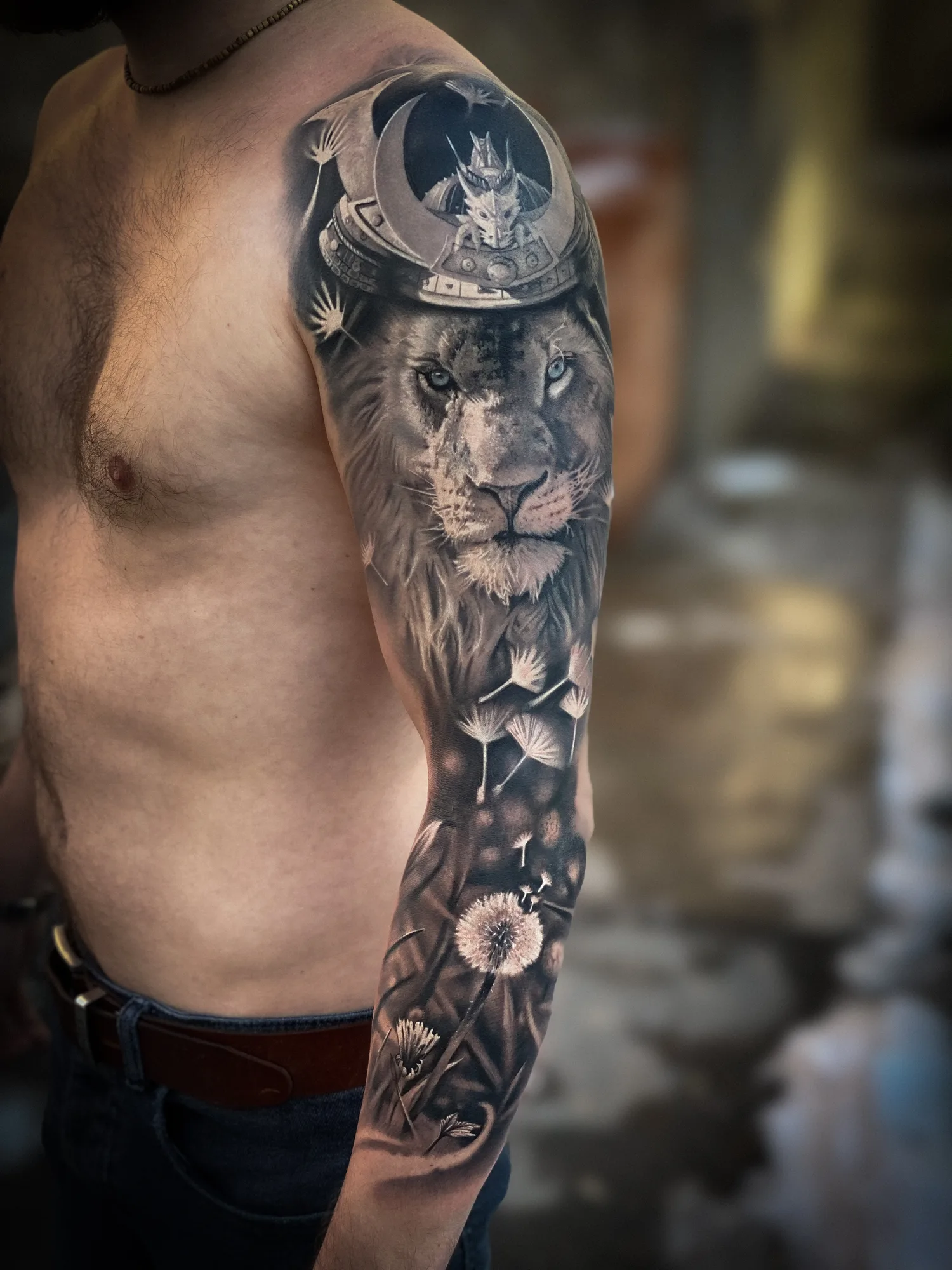 Black and grey realism full sleeve: a lion wearing a samurai kabuto helmet on the shoulder, with dandelion seeds drifting down the arm toward the wrist.