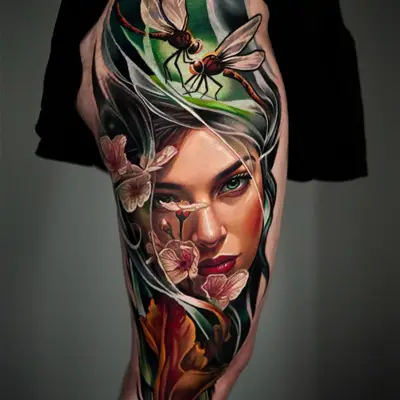 Color realism tattoo of a woman's face behind green leaves and pale blossoms with two dragonflies, on the thigh