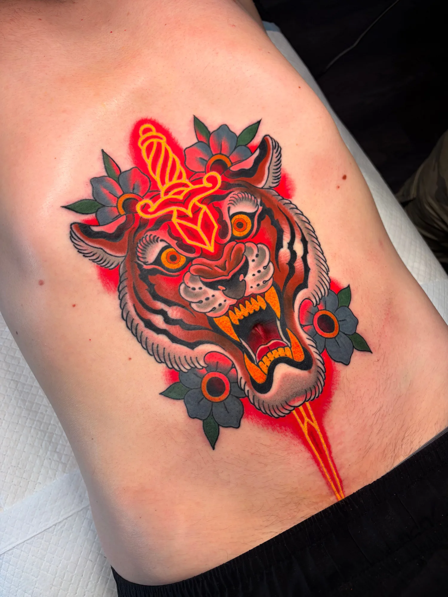 Tattoo of a tiger in an American traditional style with a dagger in neon through its head.