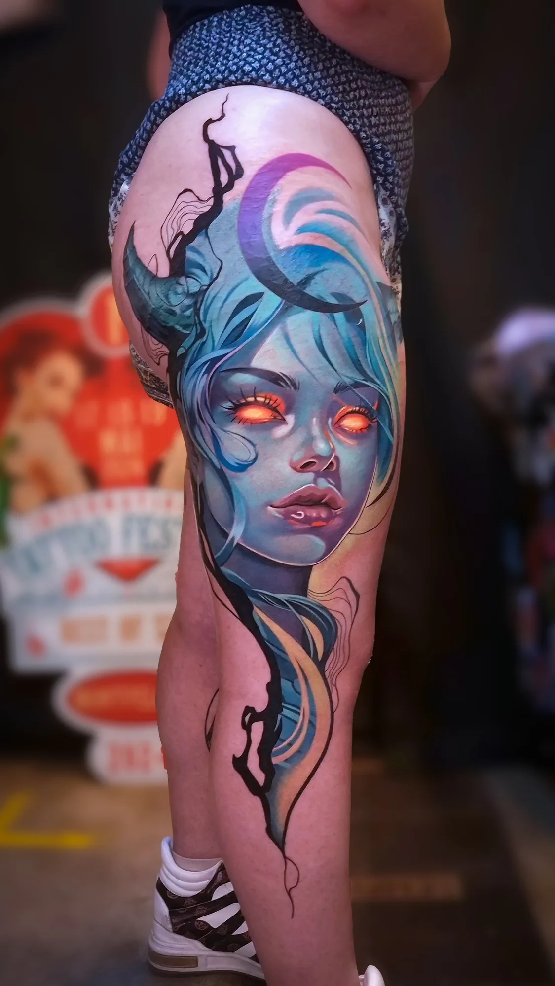 Colorful tattoo of a woman's face with blue hair and orange glowing eyes on the thigh.