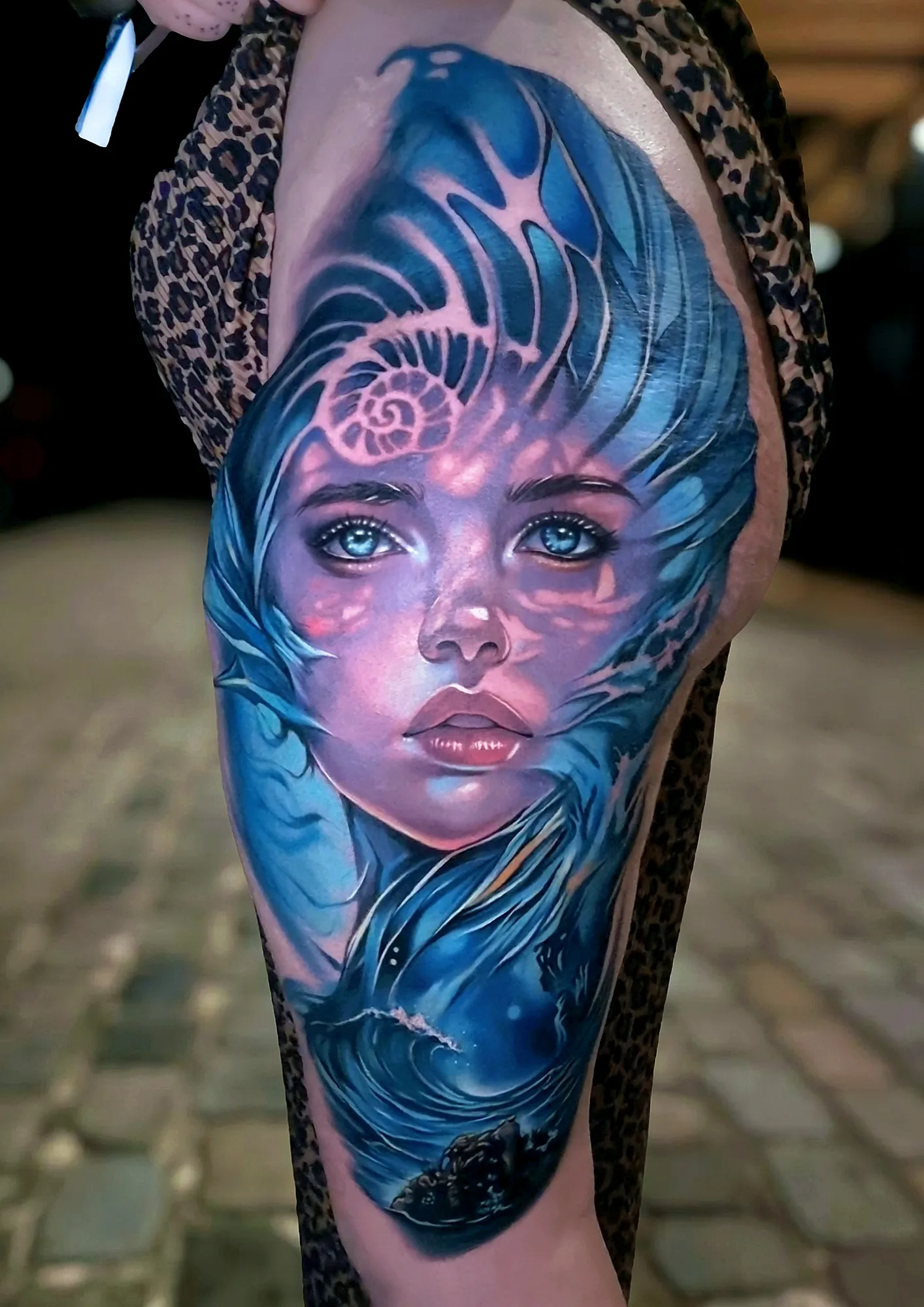 Colorful tattoo of a woman's face surrounded by waves and shells on thigh.