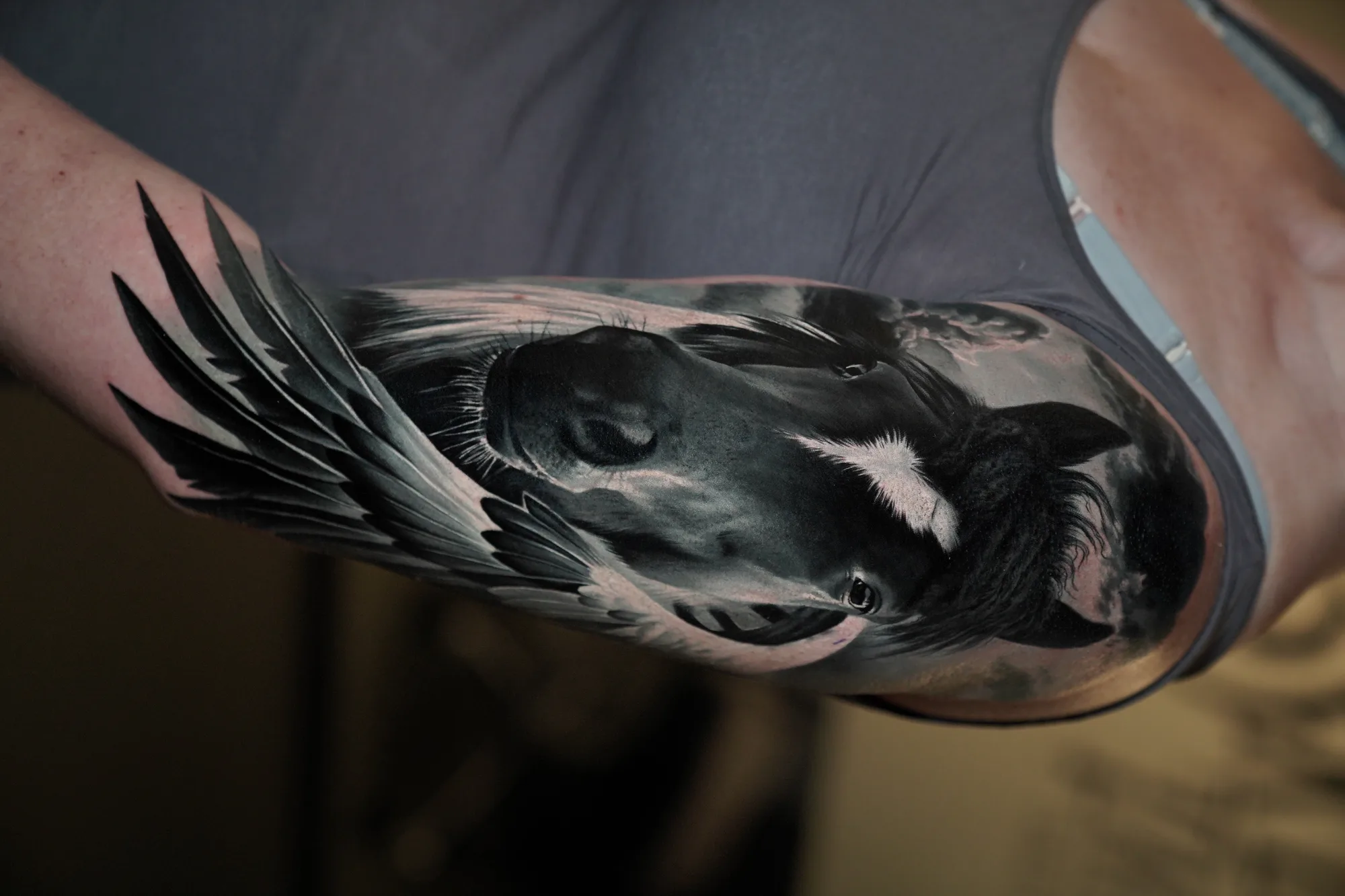 Black and grey realism tattoo on an upper arm of a dark horse's head with a white blaze, framed by a large feathered wing against a stormy sky