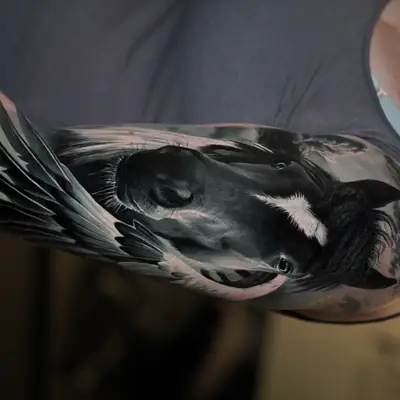 Black and grey realism tattoo on an upper arm of a dark horse's head with a white blaze, framed by a large feathered wing against a stormy sky